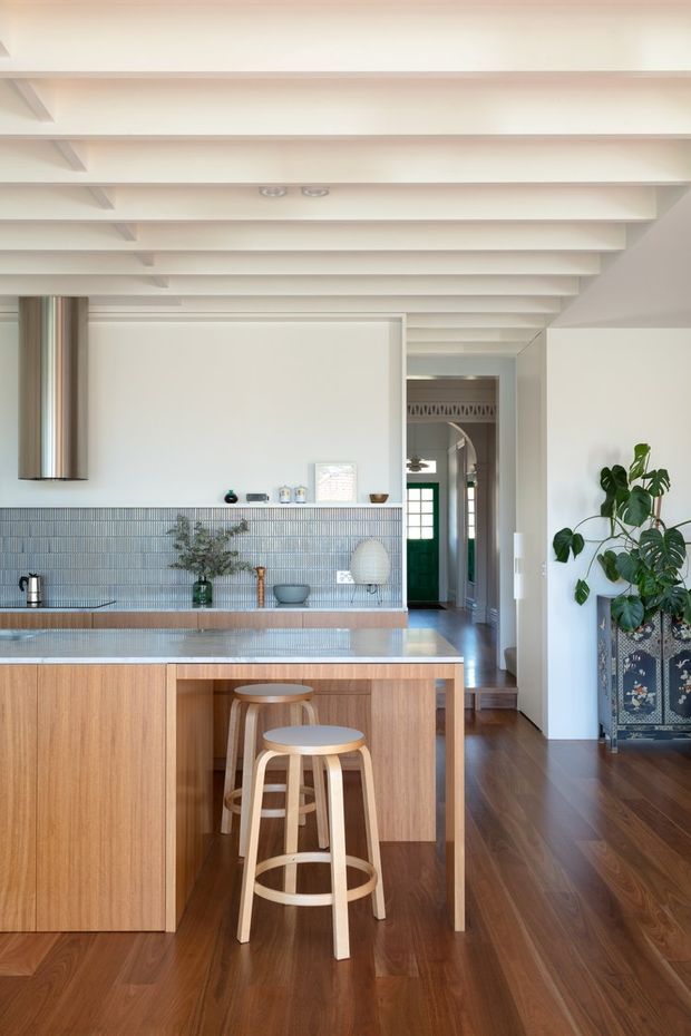 Bright contemporary kitchen featuring a timber island with waterfall-style benchtop, two stools, a fine blue-grey tiled splashback, integrated cabinetry, stainless steel rangehood, and warm timber flooring. The room has white walls and a distinctive white painted exposed-beam ceiling. A hallway and leafy indoor plant are visible beyond the kitchen.