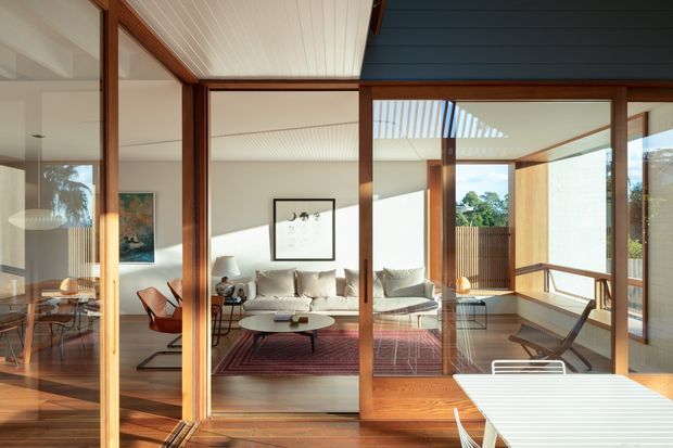 A warm, light-filled modern living area viewed through extensive timber-framed glazing and sliding doors. The room contains a pale sofa, sculptural lounge chairs, a low oval coffee table, artwork and a patterned rug, with an adjacent dining area visible to the left and an outdoor or terrace setting visible beyond the glazing. The interior features timber flooring, exposed timber framing, a white slatted ceiling, and strong indoor-outdoor connection.