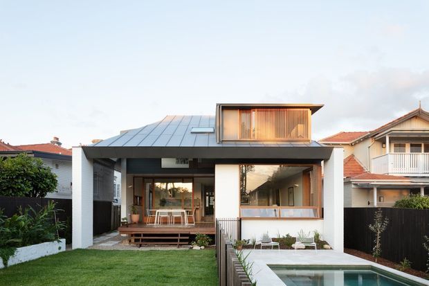 Front exterior view of a contemporary residential extension or remodel, featuring a broad low-pitched metal roof, deep roof overhang, dark structural framing, extensive glazing, and a raised timber deck opening onto a lawn. A small upper-level box with vertical timber screening projects above the main roof. The home is situated among neighbouring suburban houses and includes a compact swimming pool in the foreground.