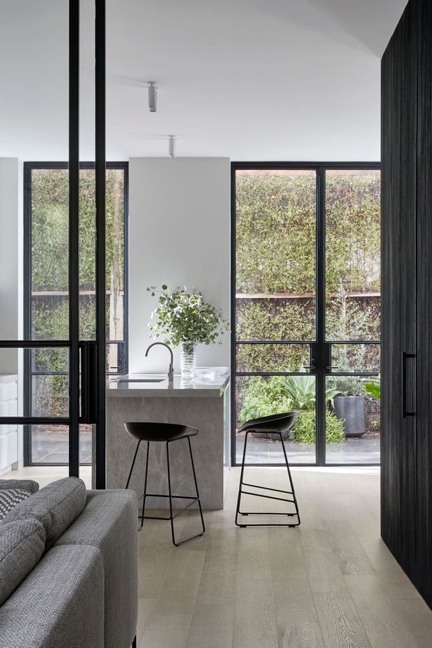 A bright, minimalist open-plan interior featuring a compact stone island with a curved black tap and two black bar stools. Floor-to-ceiling black-framed glazing looks onto a planted courtyard, while a grey upholstered sofa occupies the foreground and a dark vertical slatted feature wall frames the right side. Pale timber flooring and white walls create a clean, refined atmosphere.