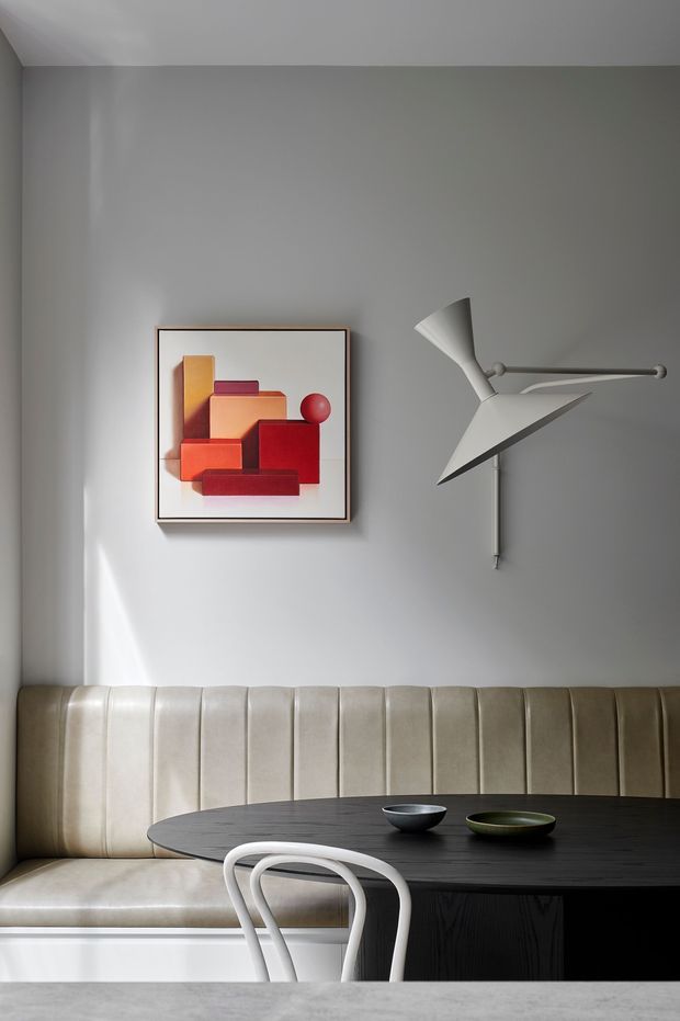 A refined dining nook with a built-in vertically channelled cream upholstered banquette wrapping around a dark round dining table. A sculptural white wall-mounted lamp and framed geometric artwork add strong mid-century-inspired accents against a warm grey wall, with daylight creating soft shadows.