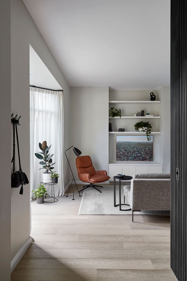 A refined contemporary living room viewed from an entry or hallway, featuring pale timber flooring, light neutral walls, sheer full-height curtains, a rust-brown leather lounge chair, grey sofa, black side tables and floor lamp, indoor plants, and built-in white shelving surrounding a television. A dark vertical slatted partition frames the right side of the image.