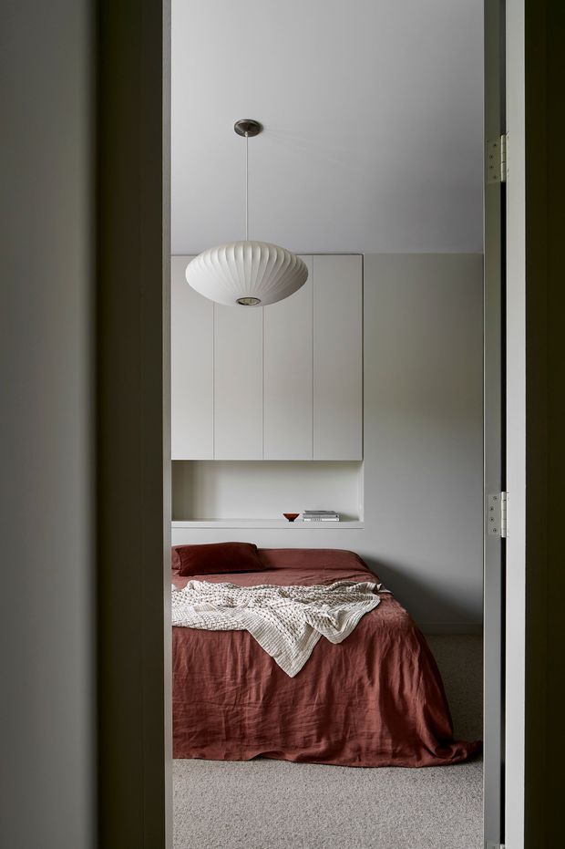 A calm, minimalist bedroom viewed through an open doorway. The room features a low bed dressed in muted rust-red linen, a textured cream throw, pale grey walls, built-in white wall cabinetry with an open display niche, and a sculptural white pendant light. Soft carpet and restrained detailing create a quiet, refined interior.