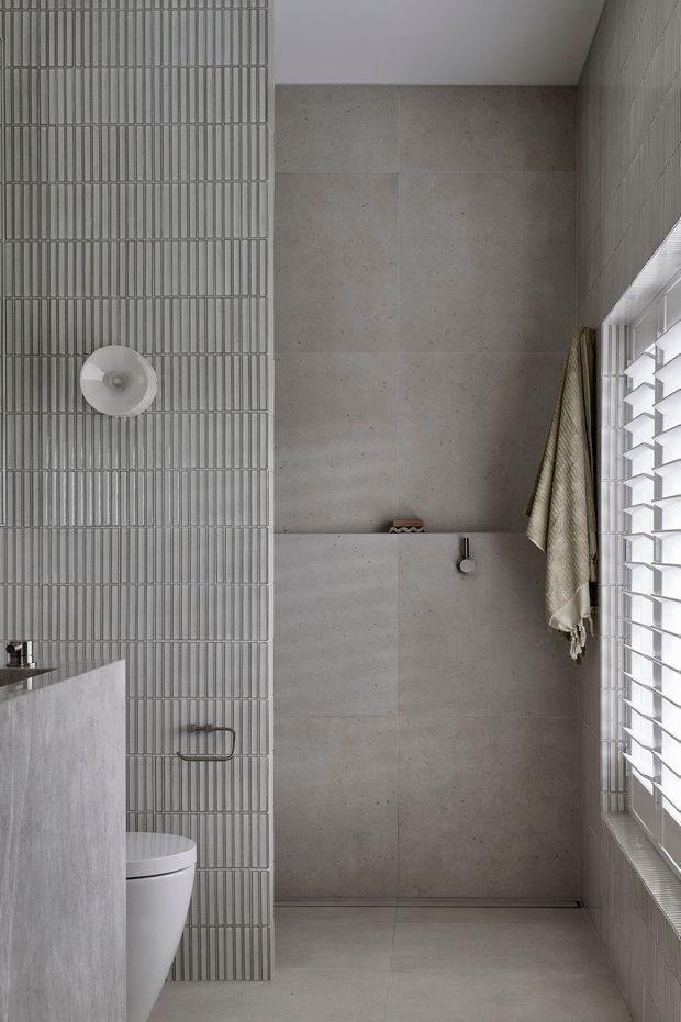 A narrow, contemporary bathroom with a walk-in shower at the rear. The space features pale grey large-format wall and floor tiles, a vertically ribbed grey tile feature wall, a white toilet partially screened by a pale stone-look vanity, brushed metal hardware, and a large louvred window admitting soft natural light. A neutral textured towel hangs beside the shower.