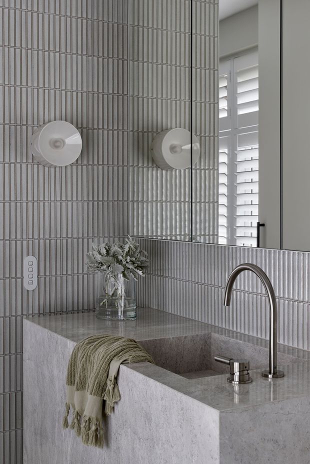 A refined contemporary bathroom vanity with a deep integrated rectangular basin in a pale grey stone surface. The walls and backsplash are clad in narrow vertically arranged grey tiles, complemented by a large mirror, round wall sconces, and white plantation shutters. A tall curved chrome tap and smaller mixer sit beside a textured neutral throw and a glass vase of foliage. The palette is monochromatic grey and white with subtle warm textile accents.