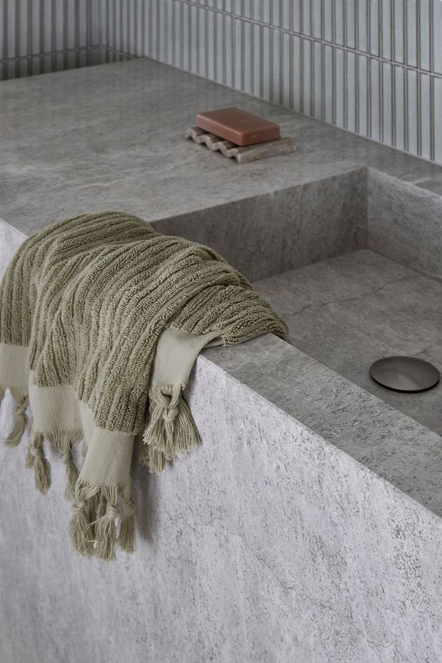 Close-up view of a minimalist bathroom featuring a deep, integrated rectangular concrete or stone bathtub with matching ledge and surround. The space is finished in grey textured stone, with narrow vertical wall tiles, a folded neutral fringed towel draped over the tub edge, a soap bar on a small tray, and a dark round drain cover. The composition is calm, tactile, and monochromatic.