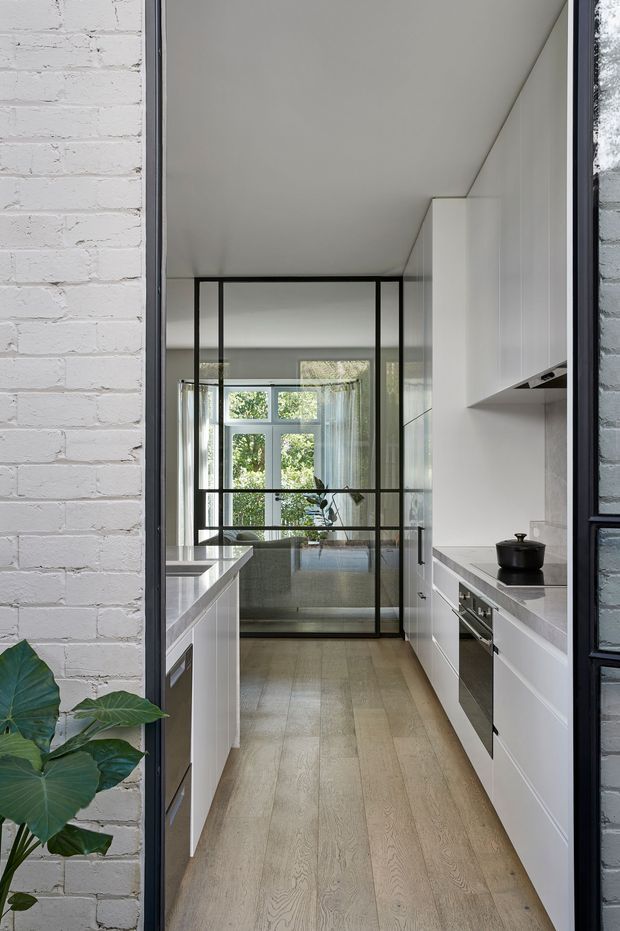 A narrow, light-filled contemporary kitchen viewed through a doorway. The galley layout has white full-height cabinetry, a pale stone benchtop, integrated stainless-steel appliances, and light timber flooring. A black-framed internal glazed partition at the far end opens toward a bright adjoining room, while a white-painted brick wall and leafy plant frame the entrance.