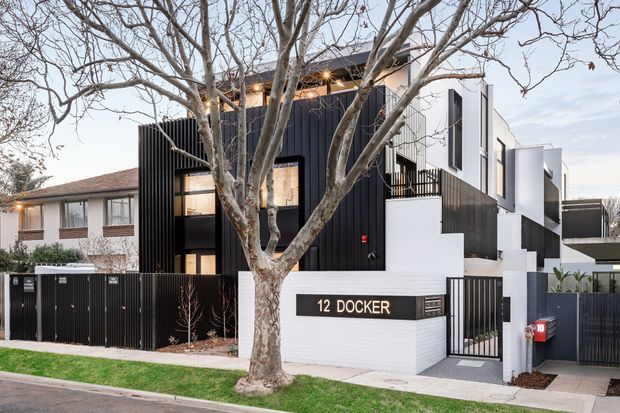 Street-facing view of a contemporary multi-unit residential development with a bold black-and-white facade, vertical dark cladding, projecting box forms, balconies, front fencing, and a landscaped sidewalk. A mature deciduous tree partially screens the building, and the entrance wall is marked “12 DOCKER”.