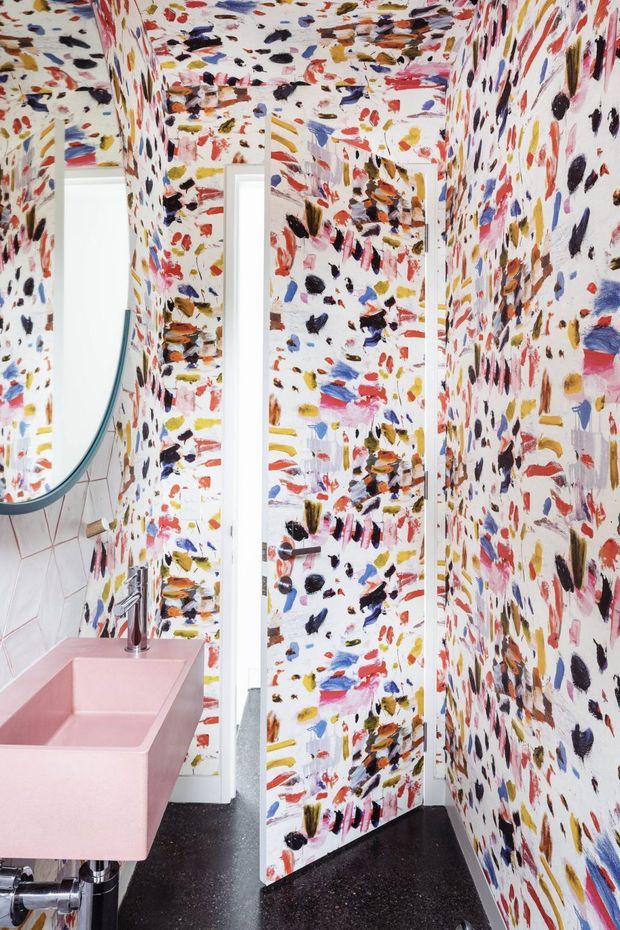 A compact, highly expressive bathroom featuring walls and ceiling covered in a multicoloured abstract brushstroke pattern. A sculptural pink wall-mounted basin with exposed chrome plumbing sits beneath a curved mirror, while a white-framed doorway and dark floor provide contrast. The space has a playful, bold, gallery-like interior treatment.