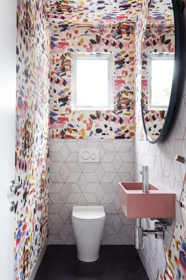 A compact contemporary powder room with a wall-mounted pink vanity, chrome tapware and exposed plumbing, a white toilet, and a large rounded mirror. The walls and ceiling feature vivid multicoloured abstract brushstroke wallpaper, while the lower walls are clad in pale hexagonal tiles with pink grout. A dark floor provides contrast.