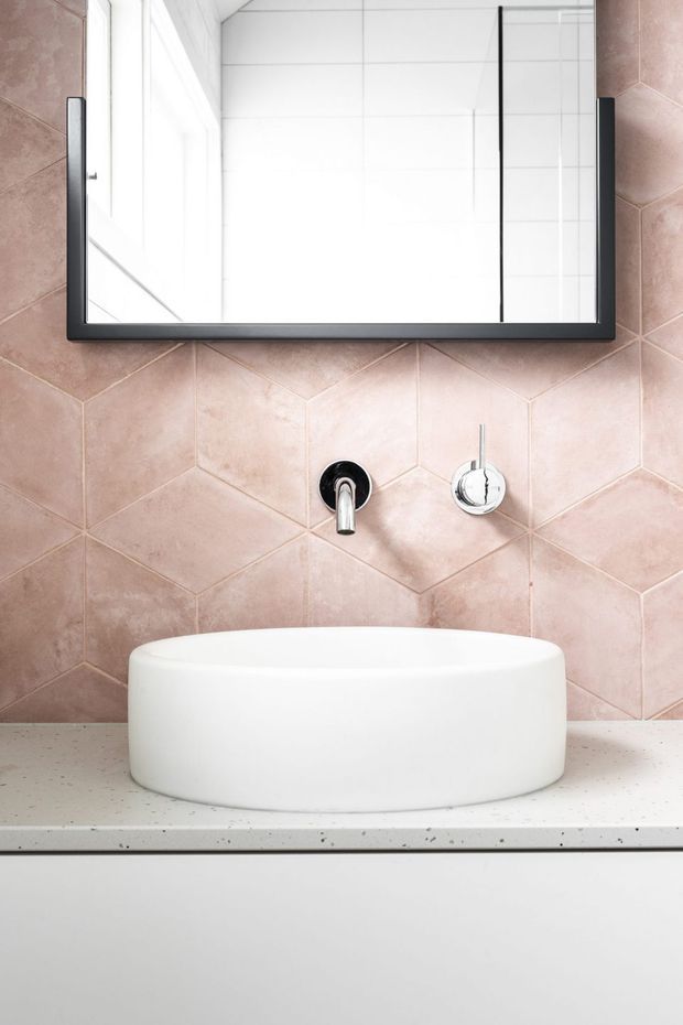 Close-up of a contemporary bathroom vanity featuring a white vessel basin on a white speckled countertop, wall-mounted chrome tapware, a large black-framed mirror, and blush-pink geometric wall tiles.