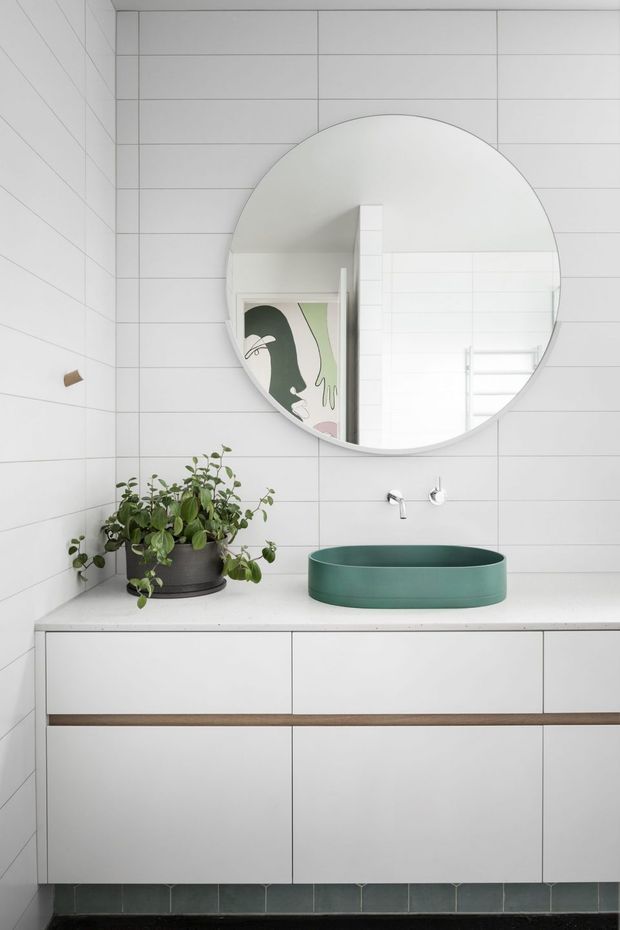 Bright, minimalist bathroom vanity with white tiled walls, a large round mirror, floating white cabinetry, a white countertop, and a distinctive deep green vessel basin. A potted plant adds greenery, while the mirror reflects a colourful artwork and a white heated towel rail.
