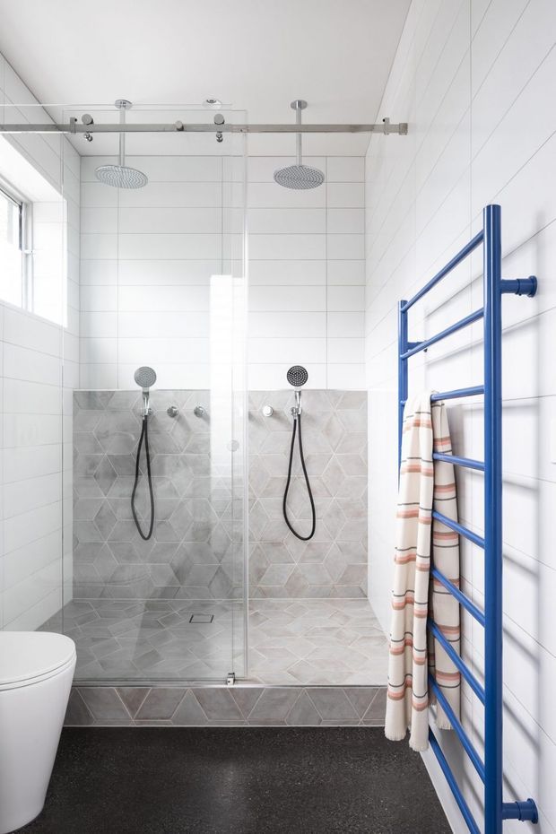 Bright contemporary bathroom featuring a spacious dual shower enclosed by clear glass, twin overhead rain showerheads, handheld shower fixtures, and grey geometric-patterned wall and floor tiles. A vivid blue heated towel rail with striped towels provides a strong accent, while a white toilet and large window complete the functional space.