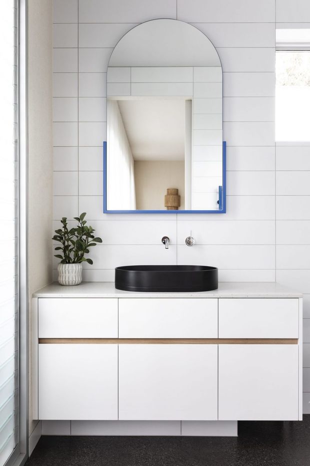 A bright contemporary bathroom vanity area featuring white tiled walls, a floating white cabinet with timber-toned trim, a matte black vessel basin, wall-mounted chrome tapware, and a distinctive arched mirror with a vivid blue frame. A small potted plant adds greenery, while a dark floor provides contrast.