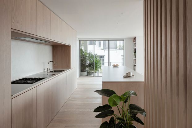 A bright, minimalist galley kitchen with pale timber cabinetry, integrated appliances, a light benchtop, and a compact island or dining counter. Vertical timber battens form a feature wall on the right, while full-height glazing at the rear brings in daylight and overlooks greenery and neighbouring buildings. A potted plant adds an organic accent to the otherwise restrained interior.