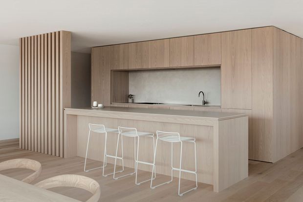 Minimalist open-plan kitchen with extensive light timber cabinetry, a large island with integrated seating, and a recessed sink niche framed by full-height joinery. Vertical timber slats partially screen the space, while pale wood flooring and white bar stools create a calm, cohesive interior.