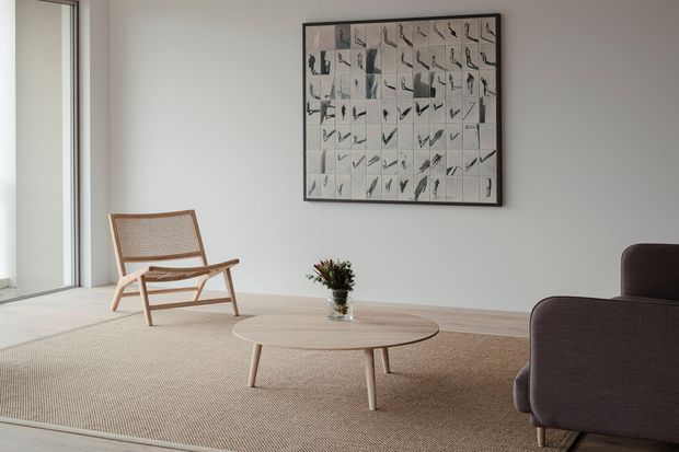 A minimalist living room with pale walls, expansive glazing at left, a low round timber coffee table, woven neutral rug, cane-and-timber lounge chair, and dark upholstered armchairs. A large monochrome framed artwork hangs above the seating area, with a small vase on the coffee table. The space is softly lit, calm, and sparsely furnished.