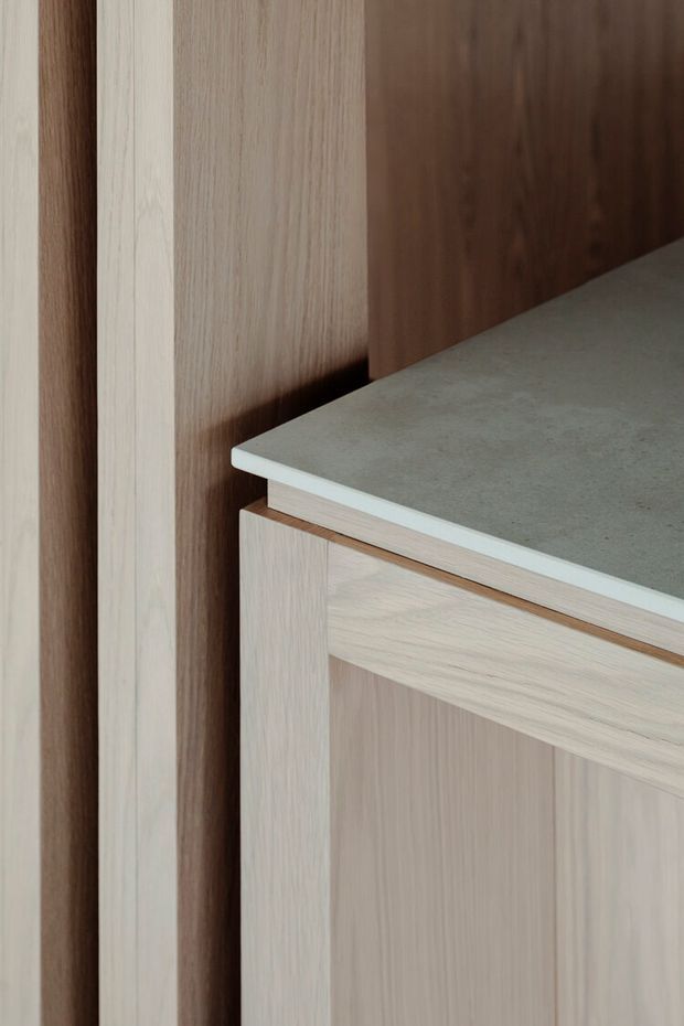 Close-up detail of pale timber-grain built-in cabinetry or joinery beside a darker timber panel, with a grey stone-look benchtop featuring a thin light edge. The image focuses on material transitions, cabinet framing and refined minimalist detailing.