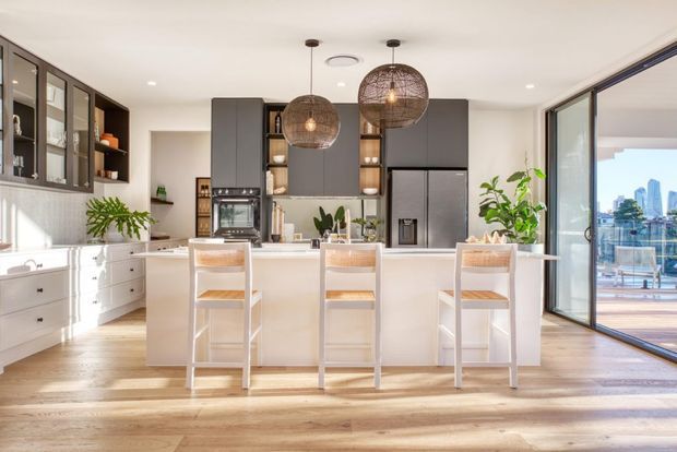 Bright contemporary open-plan kitchen with a large white island and three stools, dark grey overhead cabinetry, integrated appliances, light timber flooring, pendant lights, and expansive glazed doors opening to a balcony or terrace with an urban waterfront view. Indoor plants and warm natural light add a relaxed, inviting feel.