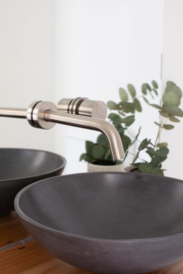 Close-up of a contemporary bathroom vanity featuring a large round dark grey vessel basin on a timber benchtop, paired with a wall-mounted brushed metal faucet. A second basin and a eucalyptus plant are visible in the background against a white wall.