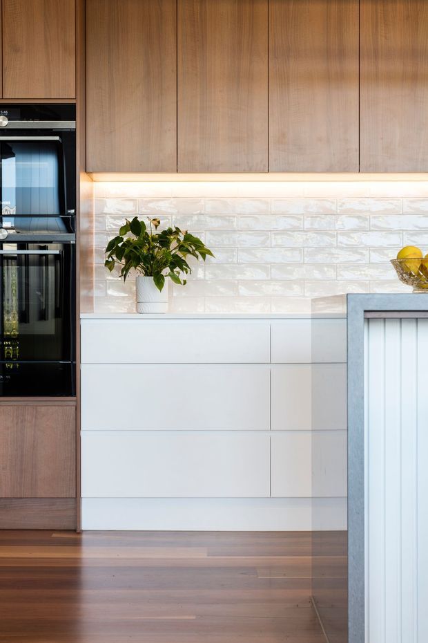 Close-up view of a contemporary kitchen featuring warm timber overhead cabinetry and integrated black appliances, white handleless lower cabinetry, a glossy white brick-pattern tiled splashback with under-cabinet lighting, and timber flooring. A plant and fruit bowl add small decorative touches, while the edge of a pale stone island is visible at right.