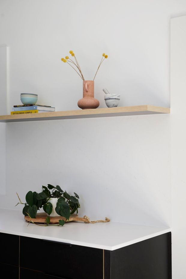 A minimalist built-in display and storage area with white walls, open light timber shelving, and a sleek black lower cabinet topped with a white surface. The shelves hold curated ceramics, books, a terracotta vase with dried stems, and a leafy potted plant. The composition is clean and contemporary with strong black-and-white contrast and natural material accents.