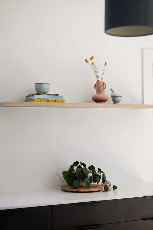 A minimalist built-in shelving and cabinetry composition. A light timber open shelf displays stacked books, a ceramic bowl, a blush-toned vase with dried stems, and a small sculptural vessel. Below, a white benchtop or surface holds a potted trailing plant above dark matte cabinetry with slim horizontal handles. A large dark pendant light is partially visible overhead, and the overall styling is clean and restrained.