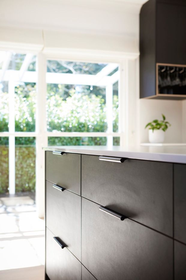 Bright contemporary kitchen detail featuring a dark charcoal built-in island or cabinetry with slim black handles, a light benchtop, white-framed windows, and a small open display shelf with a potted plant. Abundant natural light and greenery are visible beyond the glazing.