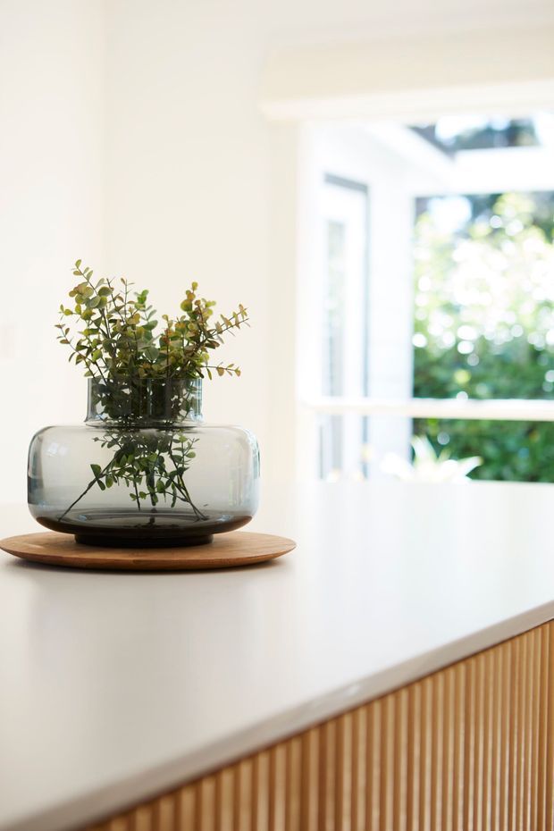 Bright close-up of a minimalist interior vignette featuring a clear glass vase with leafy greenery on a round wooden tray, placed on a white countertop. A large bright window is visible in the background, while the foreground includes a fixed vertical timber-slat cabinet or island front.