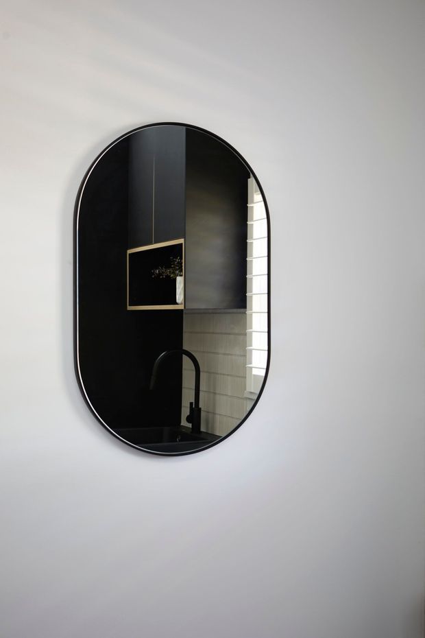 A vertically oriented, rounded-arch mirror with a slim black frame is mounted on a white wall. Its reflection shows a refined dark bathroom vanity area with black cabinetry, a black curved faucet, a dark benchtop, pale vertically textured wall tiles, a narrow louvred window, and a small framed artwork with a brass-toned frame.