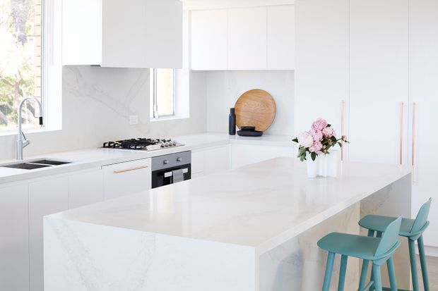 Bright, minimalist white kitchen with a large waterfall-edge island, integrated white cabinetry, pale veined stone surfaces, stainless-steel appliances, and a gas cooktop. Two teal bar stools and a vase of pink flowers provide colour accents. Large windows bring in abundant natural light.
