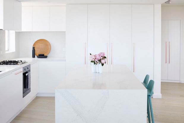 Bright, clean contemporary white kitchen with a large white stone island, integrated full-height cabinetry, pale timber flooring, slim pale-wood handles, and teal bar stools. A gas cooktop and built-in oven sit along the left wall, while a small floral arrangement and round timber board provide restrained accents.