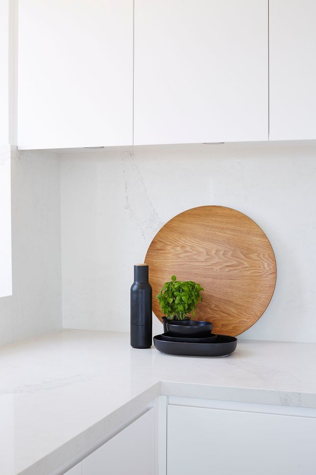 Bright minimalist kitchen corner with white flat-panel overhead and base cabinetry, pale veined stone benchtops and matching splashback. A round timber serving board, black bottle, black bowls and a small green plant provide restrained styling accents.