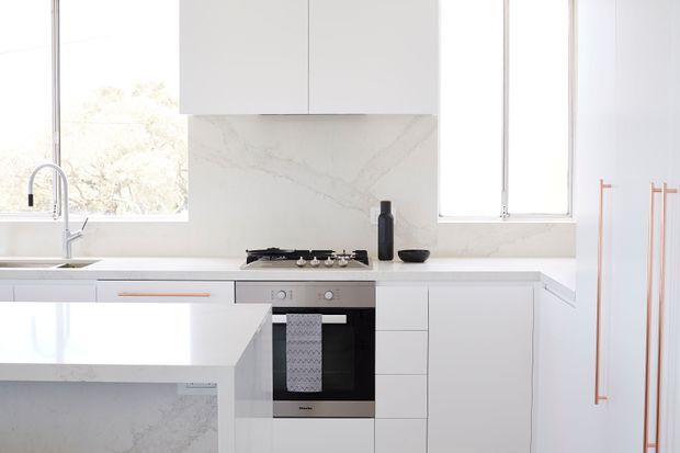 Bright minimalist contemporary kitchen with white flat-panel cabinetry, a pale veined stone backsplash and matching waterfall-edge island, integrated stainless-steel oven and cooktop, and white-framed windows. Copper-toned vertical handles add a restrained accent, while natural light creates an airy, clean atmosphere.