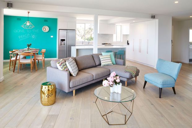 Bright open-plan living, dining and kitchen interior with pale timber flooring, white cabinetry, and large windows. A grey sectional sofa and blue accent chair surround a glass coffee table, while a turquoise feature wall defines the dining area. The room has colourful cushions, a geometric pendant light, and a fresh contemporary family-home feel.