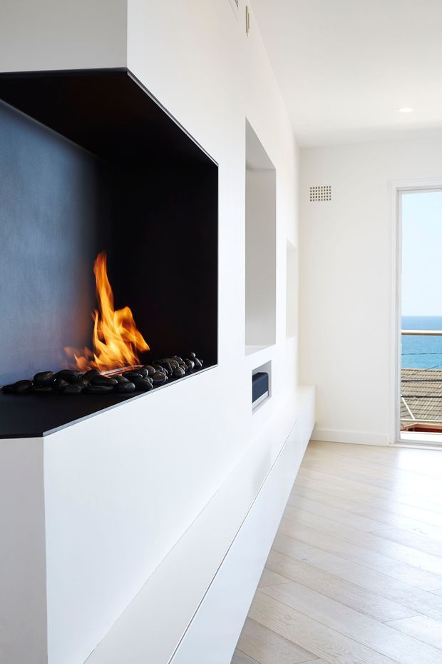 Bright minimalist interior featuring a dramatic built-in black fireplace with exposed flames and dark decorative stones, integrated into a white media and storage wall. Pale timber flooring, white walls, recessed openings, and a glazed balcony door create a clean coastal-facing living space with an ocean view.