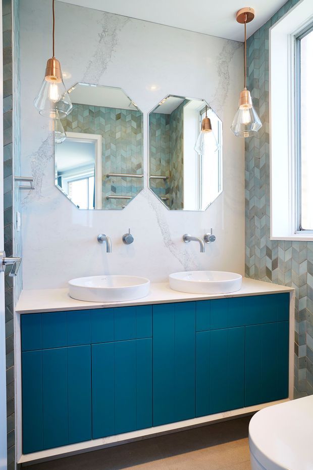Bright contemporary bathroom with a double wall-mounted vanity in deep teal cabinetry, two white vessel basins, brushed metal wall-mounted taps, and paired frameless octagonal mirrors. The walls feature large white marble-look panels and blue-green geometric patterned tiles, complemented by copper pendant lights and a large white-framed window.