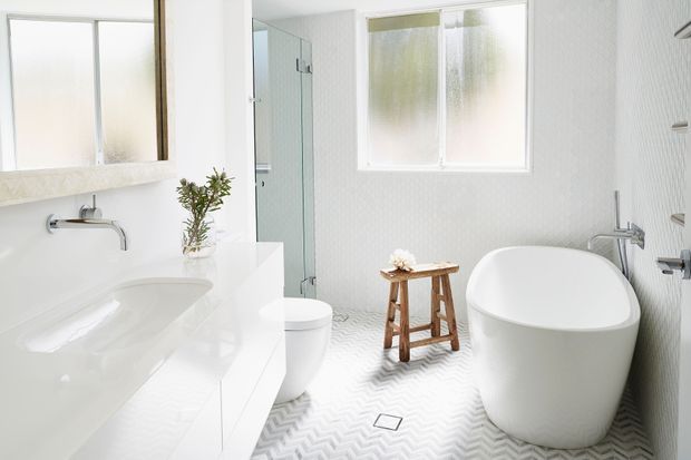 Bright contemporary bathroom with a freestanding white bathtub, wall-mounted basin and tapware, white vanity cabinetry, toilet, glass shower enclosure, frosted windows, and patterned light-grey and white floor tiles. Natural timber stool and greenery add warmth to the otherwise crisp white space.