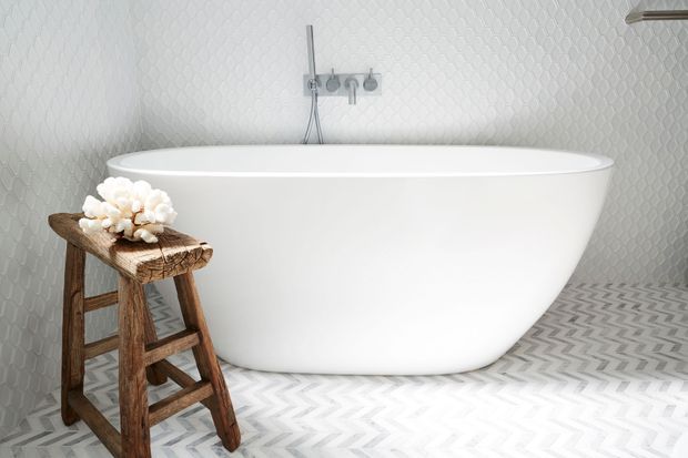 A bright, contemporary bathroom featuring a freestanding white oval bathtub against a textured pale wall. The floor is covered in a bold light-grey and white chevron pattern, with a rustic wooden stool holding white flowers beside the tub. Minimal chrome wall-mounted bath fittings and a partial towel rail are visible.
