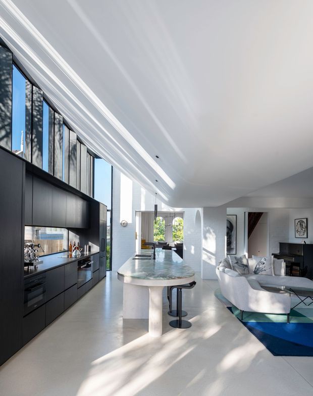 A bright, open-plan contemporary interior combining a matte black kitchen with integrated appliances, a large pale stone island with rounded edges, and an adjacent white living area. Tall floor-to-ceiling glazing and clerestory windows bring in abundant daylight, while curved white architectural walls and a sculptural ceiling transition add softness. The living room includes a white sofa, patterned cushions, a black piano, and a blue geometric rug.