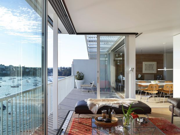 A bright contemporary living room opening onto a waterfront balcony terrace. Full-height glazing overlooks a marina, while a pergola-covered outdoor seating area extends beyond the sliding glass doors. Inside, the room features a low dark sofa and ottoman, glass coffee table, patterned rug, timber dining area with orange chairs, and warm timber wall panelling.