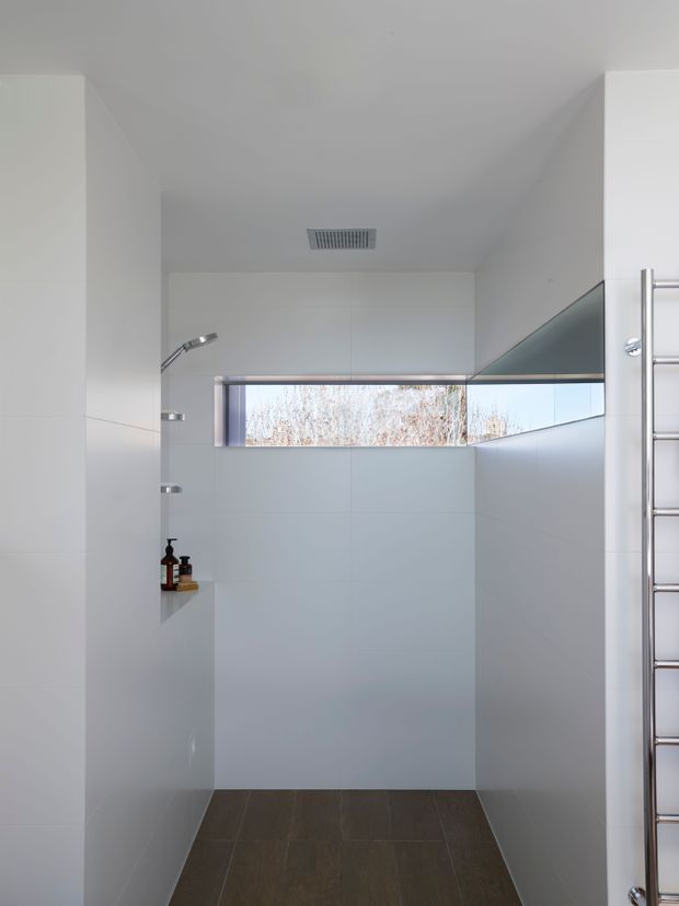Minimal, contemporary walk-in shower bathroom with white tiled walls, large dark-format floor tiles, a frameless glass shower screen, chrome shower fittings, a narrow horizontal window, recessed wall niche, and a chrome heated towel rail. The space is bright, clean, and highly pared back.