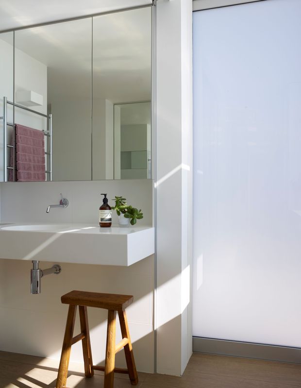 Bright minimalist bathroom with a wall-mounted white basin and chrome tapware, large mirrored cabinetry, a chrome towel rail with folded towels, and a wooden stool. A frosted glass partition or door occupies the right side, while sunlight creates strong geometric shadows across the white surfaces and timber-look floor.