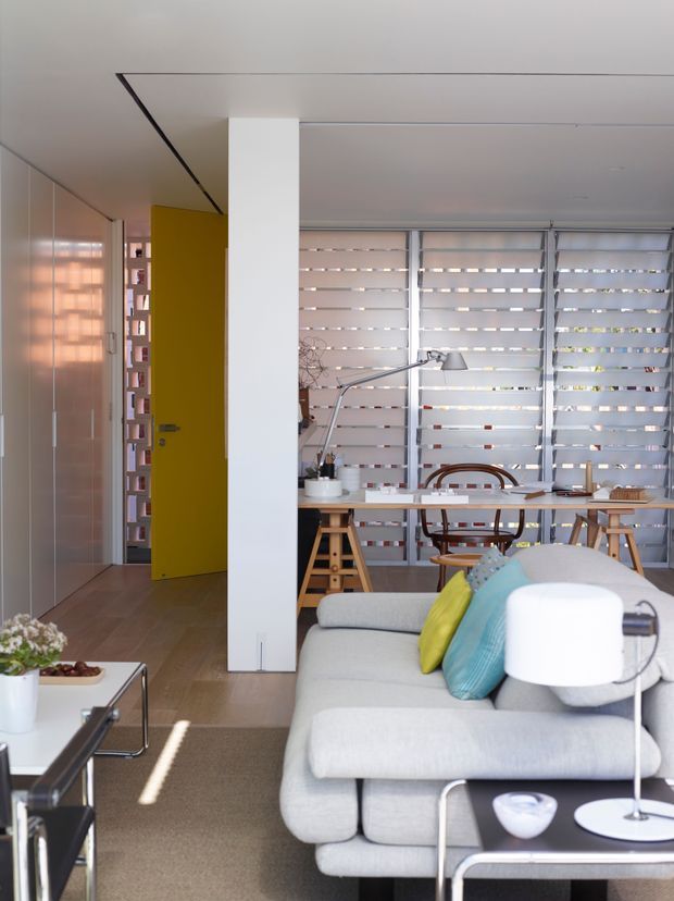 A bright contemporary open-plan living room with a grey sectional sofa in the foreground, turquoise and yellow cushions, a low black coffee table, and a compact dining/work table behind it. The room has pale timber flooring, a neutral carpeted seating area, full-height white built-in cabinetry along the left wall, a vivid yellow internal door, and broad windows fitted with horizontal translucent blinds. A white structural column divides the living and dining zones.