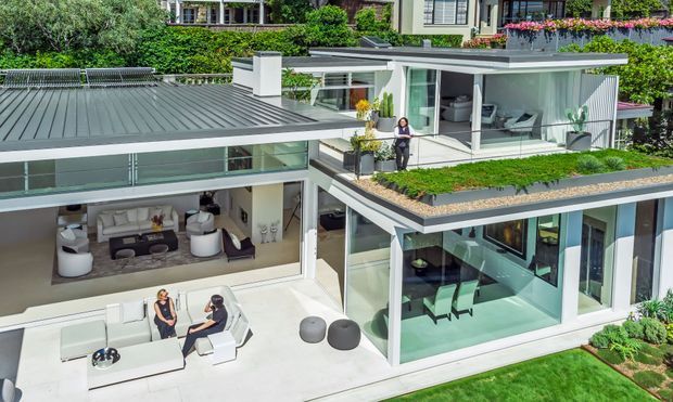 Aerial view of a contemporary multi-level residence with flat roofs, extensive glazing, large outdoor terraces, and planted green-roof areas. The home features crisp white and grey architectural surfaces, open-plan living spaces visible through the glazing, outdoor seating, and landscaped garden surrounds.