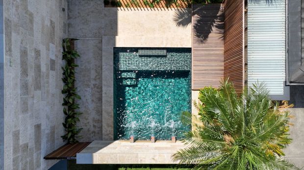 Aerial view of a compact contemporary courtyard centered on a rectangular turquoise swimming pool with water features. Pale stone paving and wall surfaces contrast with a warm timber slatted deck, linear planting, and tropical palm foliage. The composition emphasizes a private outdoor pool area and landscaped hardscape.