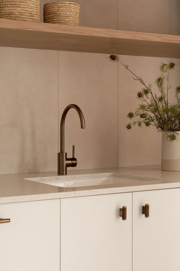 Close-up of a warm, minimalist kitchen or utility-room sink area. A bronze gooseneck tap sits above an integrated white sink set into a pale stone benchtop, with cream cabinetry and brass-toned hardware below. The backsplash is clad in vertically jointed light timber or timber-look panels, and an open timber shelf with woven baskets and a ceramic vase adds natural texture.