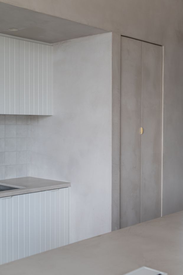A minimalist contemporary kitchen in a soft grey palette, featuring vertically panelled light cabinetry, a grey stone or concrete-look benchtop, tiled splashback, smooth plastered walls, and a tall flush concealed door with a small brass-gold handle.