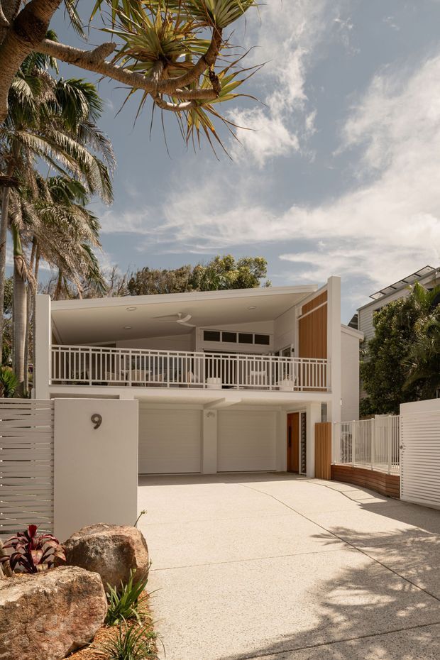 A bright tropical coastal residential facade viewed from the driveway. The two-storey home has a broad upper-level balcony with white metal balustrading, a covered outdoor area, white rendered walls, timber-look vertical cladding accents, garage doors, and a textured concrete driveway. Mature palms and tropical planting frame the property.