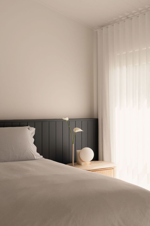 A softly lit, minimalist bedroom corner viewed from the bed. The room features a dark charcoal vertical-panel headboard or half-height wall treatment, warm off-white walls, pale neutral bedding, sheer white floor-to-ceiling curtains, and a light timber bedside table with a small spherical lamp and simple floral arrangement. Natural daylight creates a calm, refined atmosphere.